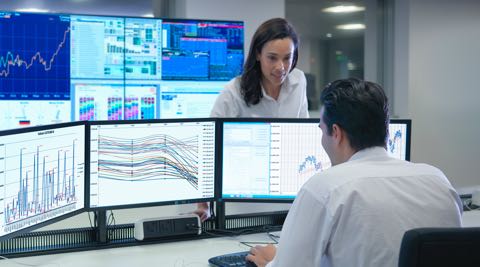 over-the-shoulder view of a man facing two large monitors while using the keyboard, and a standing woman facing him, and more monitors behind her